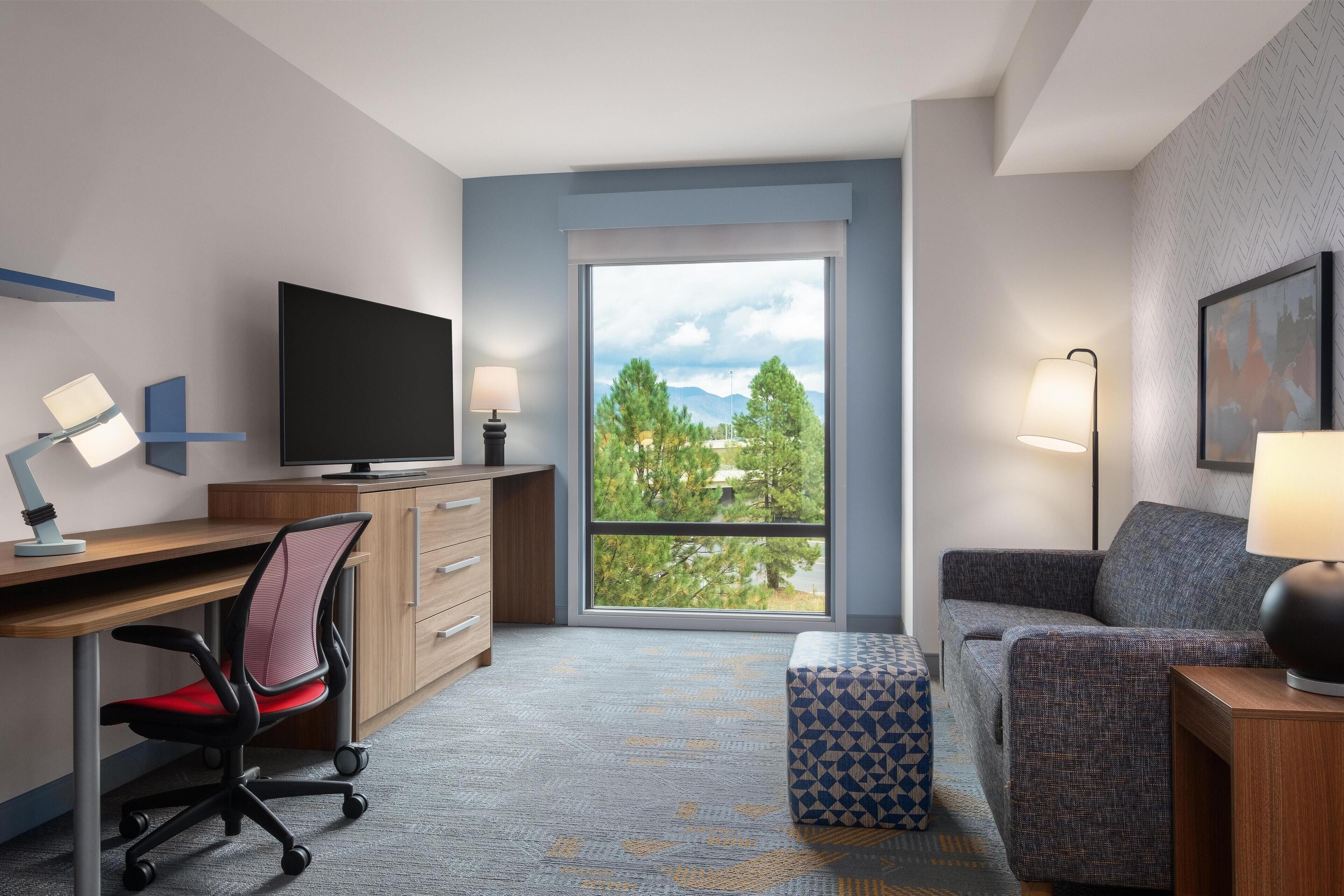 A hotel room with a desk, chair, television, sofa, and a large window with a view of trees and mountains.