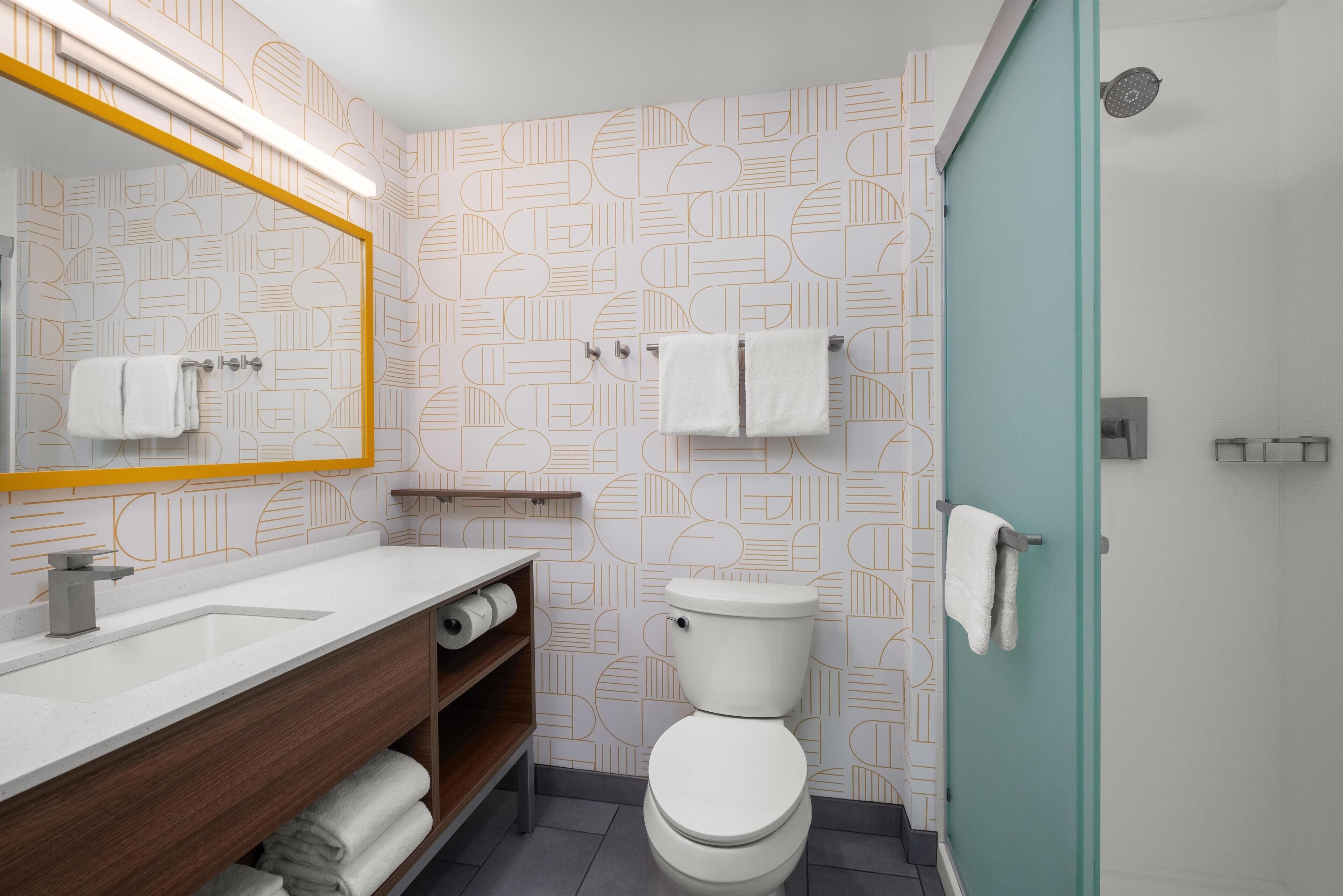 A modern bathroom with a geometric patterned wall, a white toilet, a wooden vanity with a mirror, and a shower area.