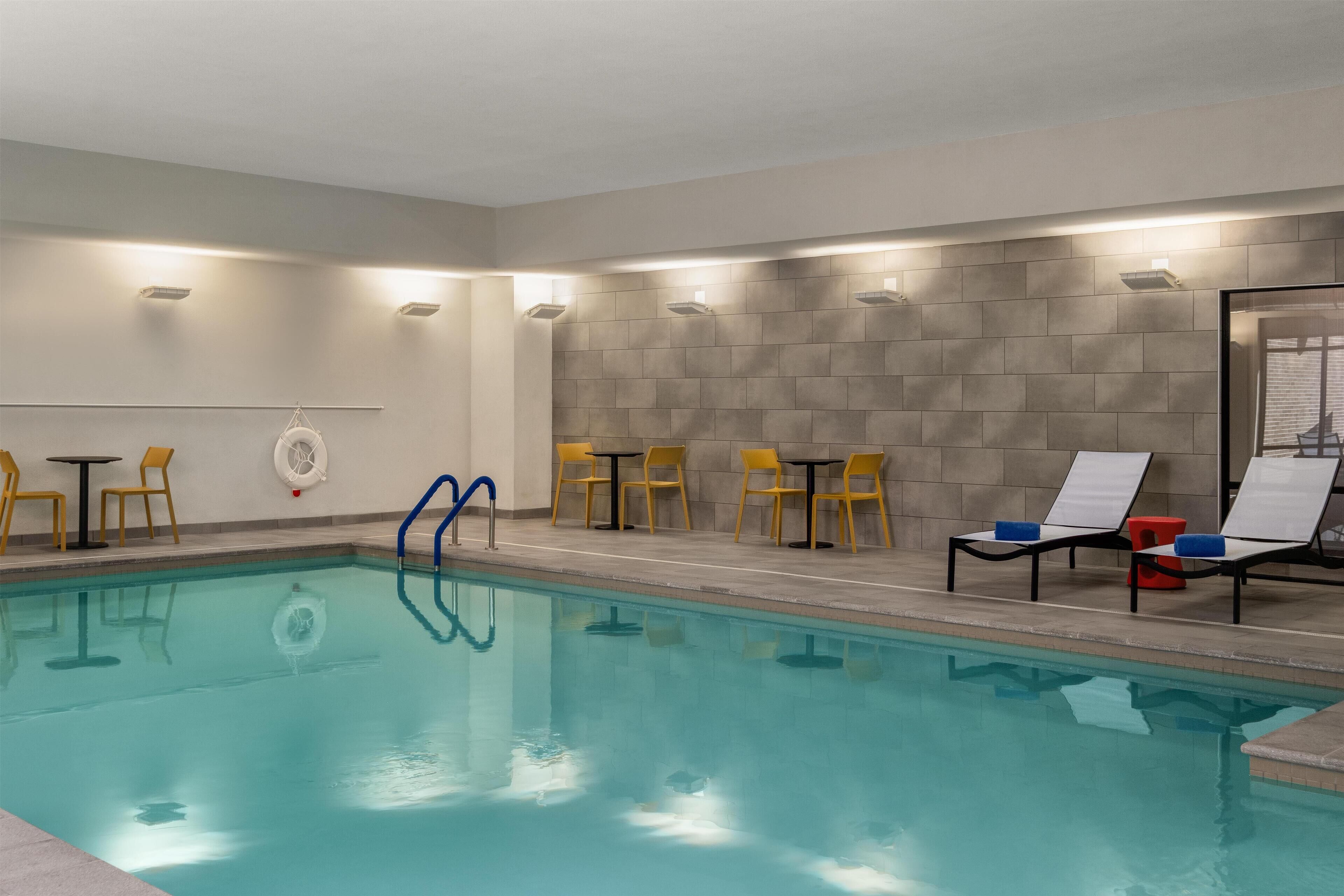 A swimming pool with clear water, surrounded by yellow chairs and tables, and a tiled wall.