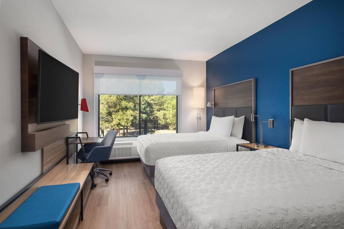 A hotel room with two beds, a desk with a chair, a TV mounted on the wall, and a window with a view of trees.