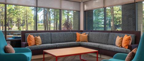 A modern interior with a sectional sofa, teal armchairs, and a wooden coffee table. Large windows offer views of trees and a brick building.