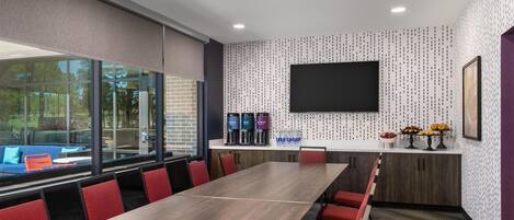 A conference room with a long table, red chairs, a large window, a flat-screen TV, and a wall-mounted coffee station.