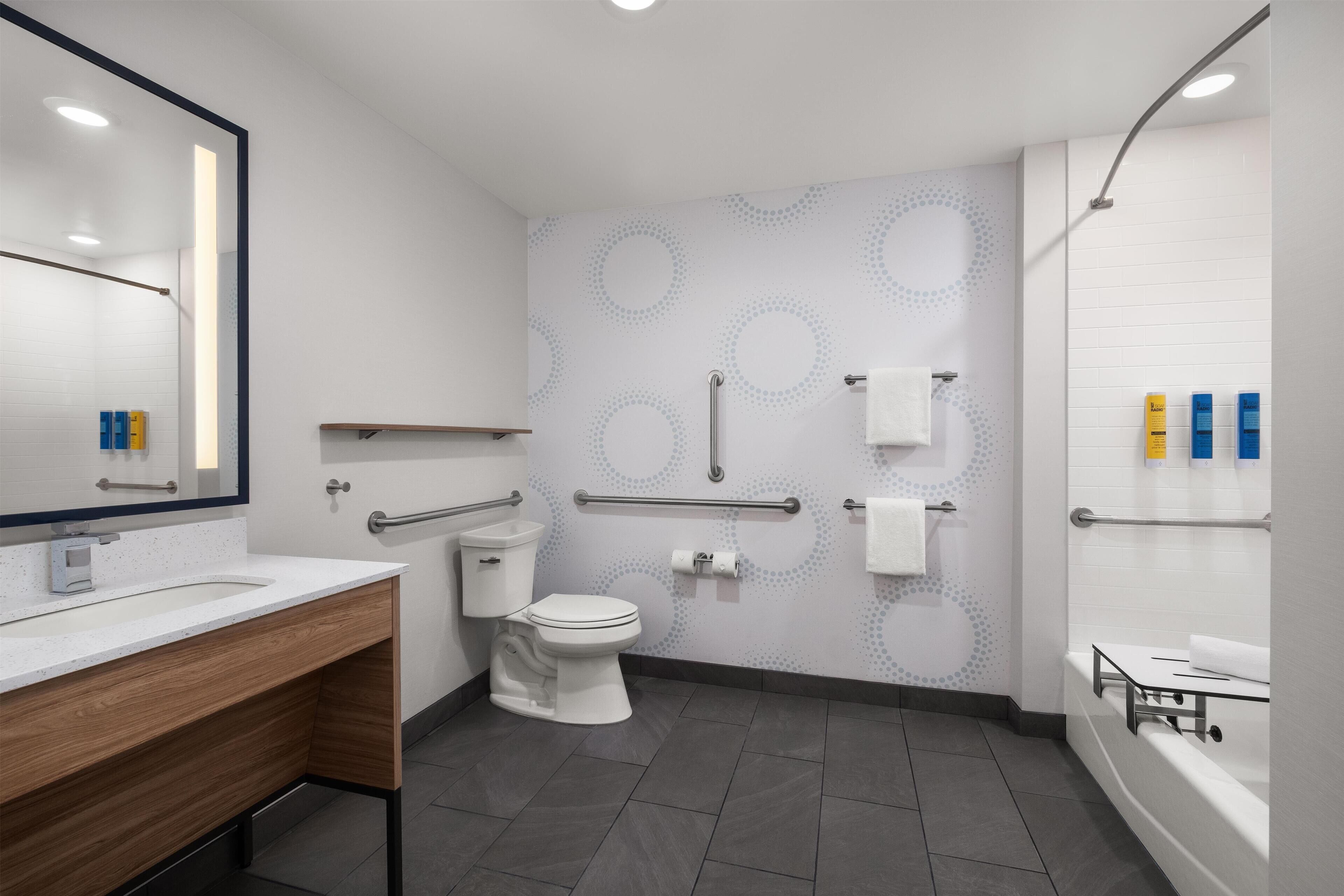 A modern bathroom with a white sink, toilet, and bathtub. It includes a mirror, towel rack, and handrails.