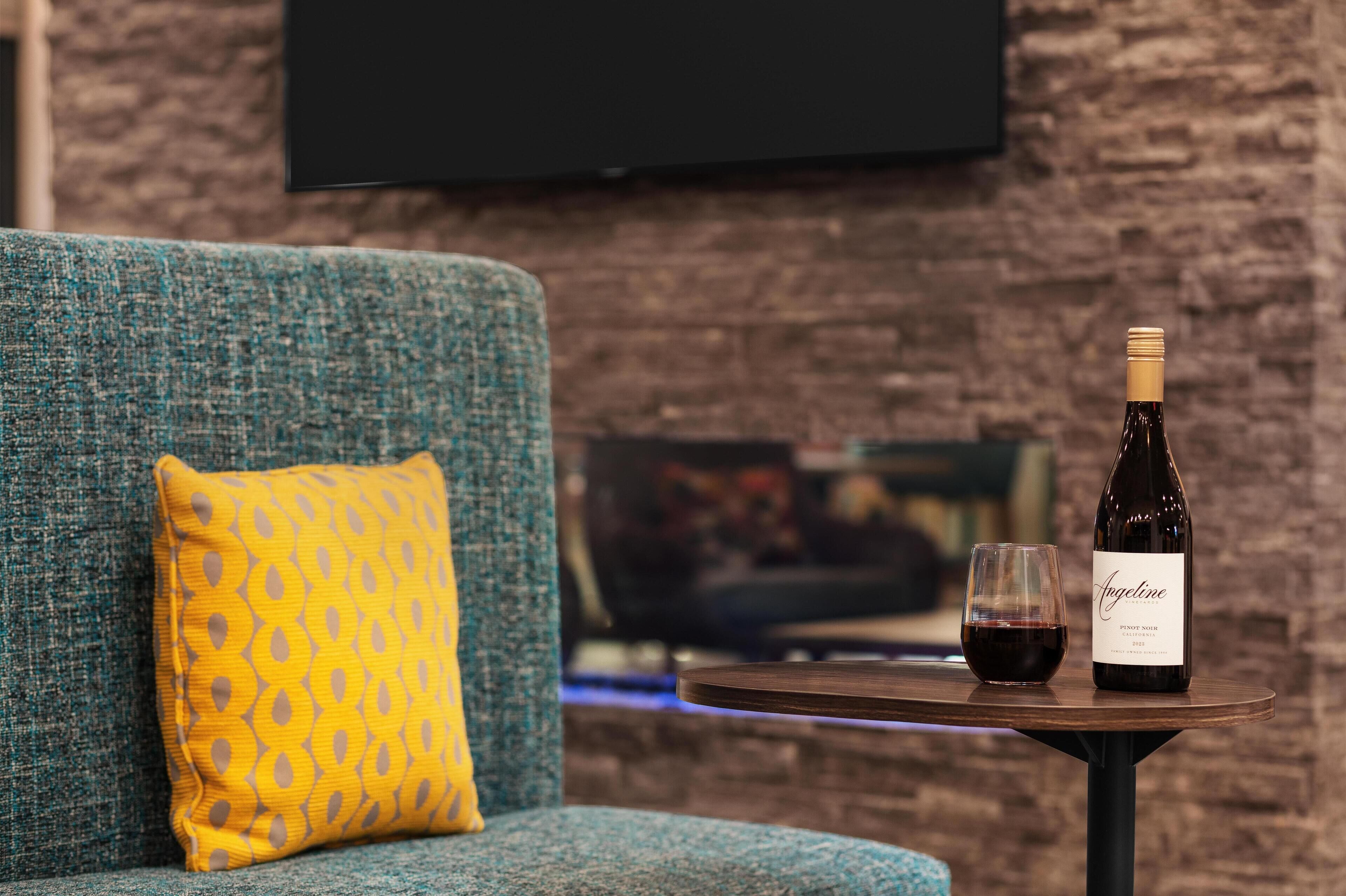 A cozy seating area with a blue sofa and a yellow patterned pillow, a small round table with a bottle of wine and a glass, and a brick wall in the background.