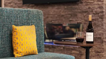 A cozy seating area with a blue sofa and a yellow patterned pillow, a small round table with a bottle of wine and a glass, and a brick wall in the background.