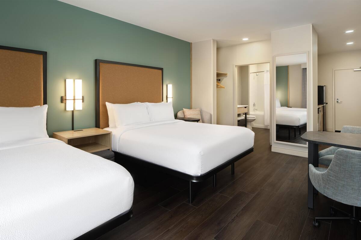 Habitación de hotel con dos camas, un escritorio, una silla, un espejo y un baño visible a través de una puerta abierta.