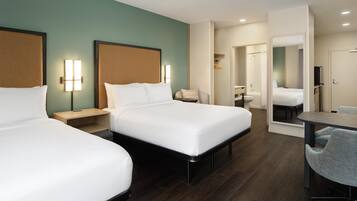 Quarto de hotel com duas camas, uma escrivaninha, uma cadeira, um espelho e um banheiro visível através de uma porta aberta.