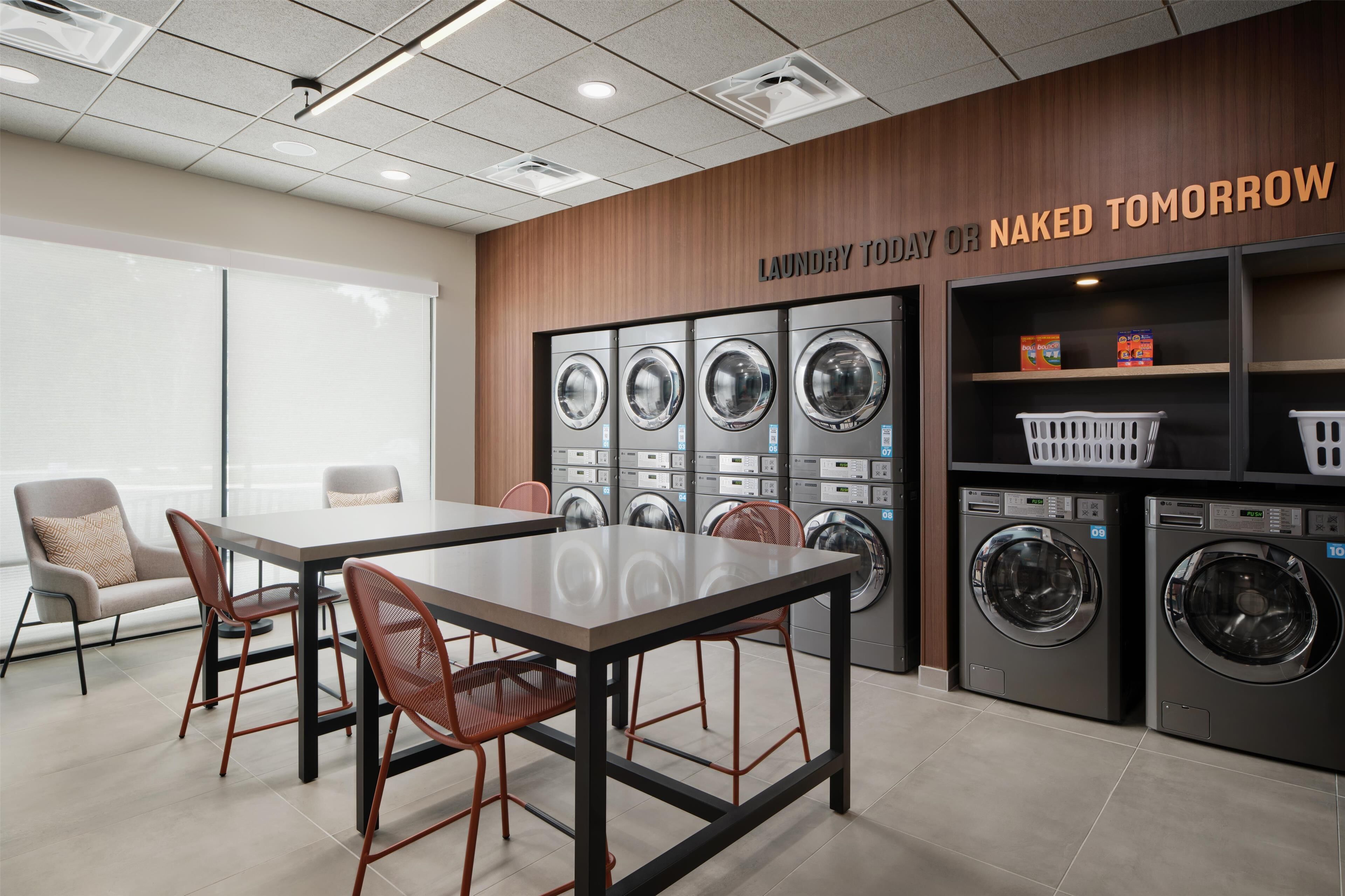 A modern laundry room with multiple washing machines, a table with chairs, and a sign that reads "Laundry Today or Naked Tomorrow".