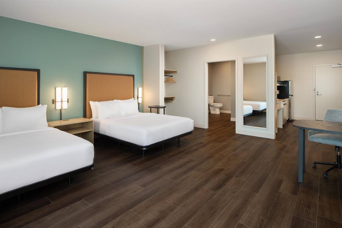 Habitación de hotel con dos camas individuales, un escritorio con silla, una mesita, un espejo y un baño visible a través de una puerta abierta.