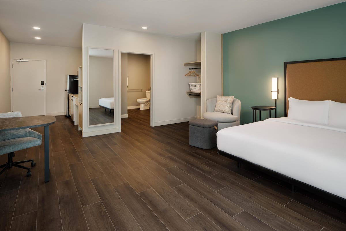 Habitación de hotel con una cama grande, un escritorio, una silla, una mesita, un sofá, un cubo de basura, un espejo, un armario y una lámpara.