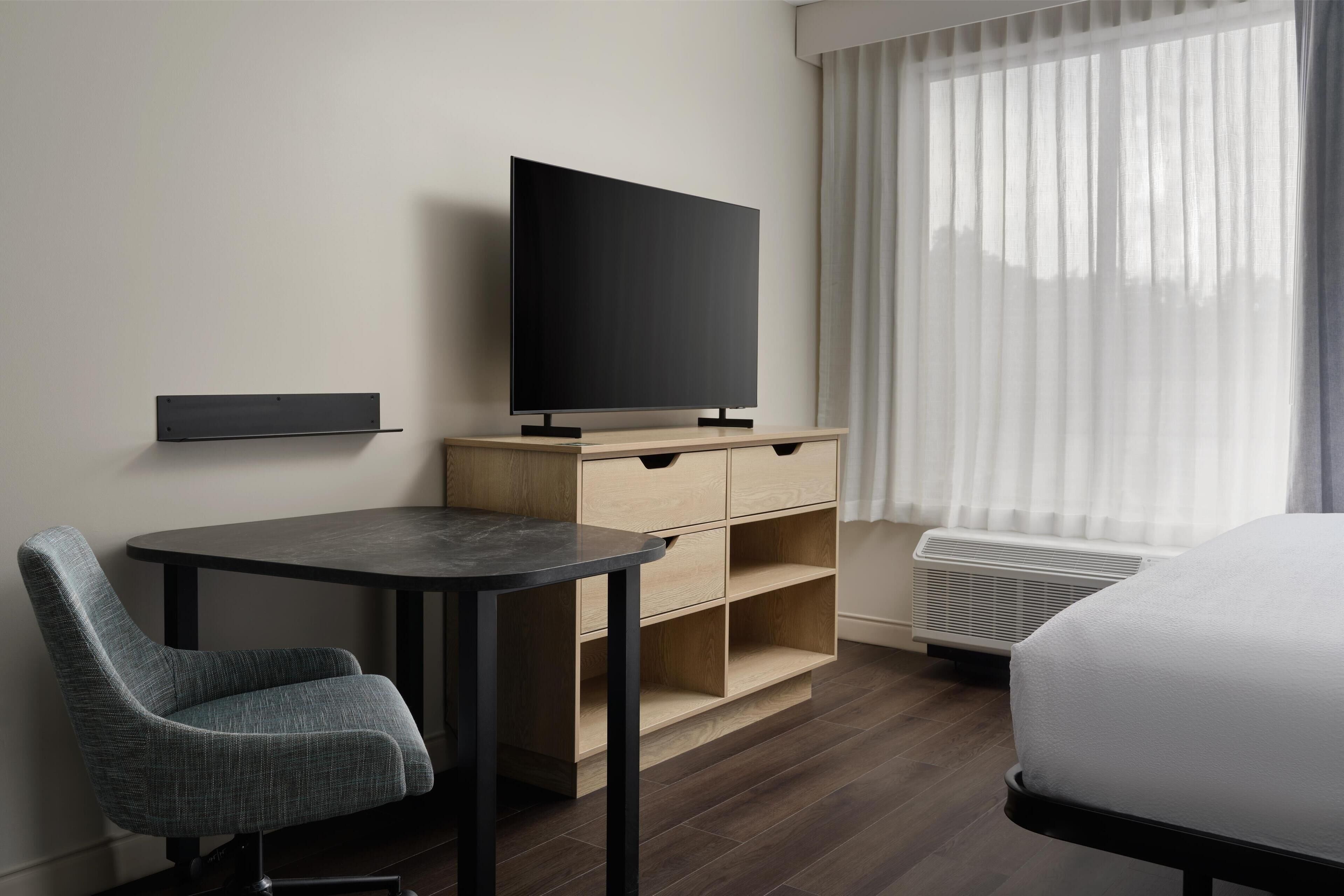 Ein Hotelzimmer mit Flachbildfernseher auf einem Holzboden, einem Schreibtisch mit Stuhl, einem Bett und einem Fenster mit transparenten Vorhängen.