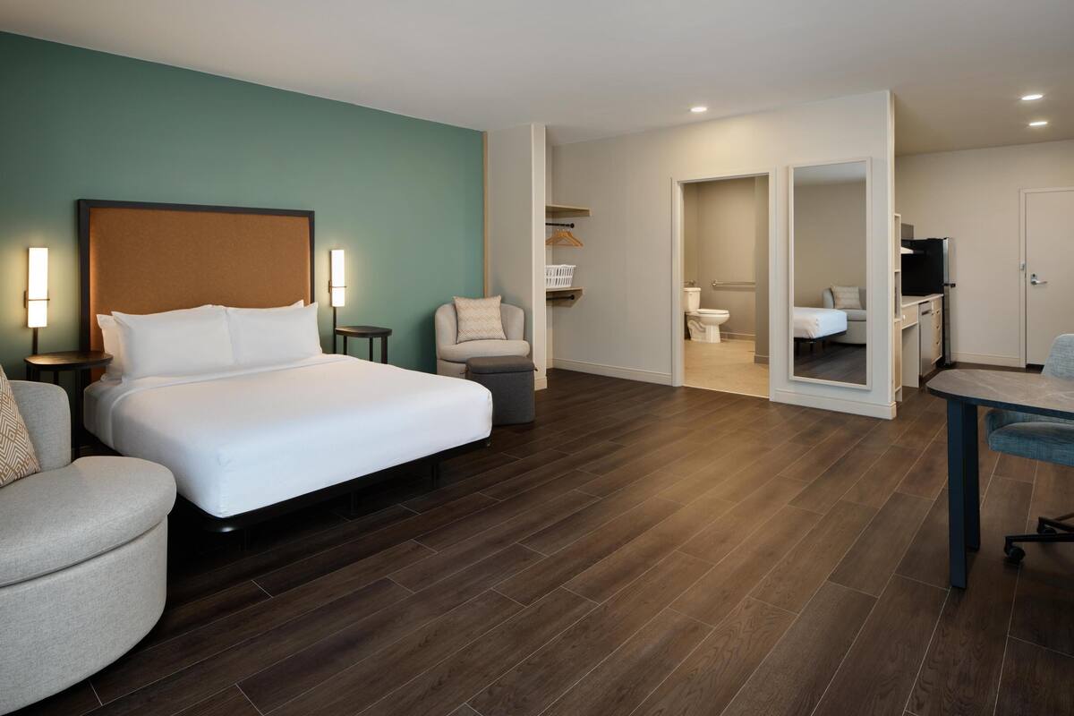 Habitación de hotel con una cama grande, una silla, un escritorio y un baño visible a través de una puerta abierta.