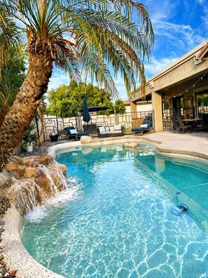 A swimming pool with a waterfall feature, surrounded by lounge chairs and a palm tree. - Luxe Retreat- Private Pool -BBQ- Peaceful Retreat- Comfort-Relax-Prime Location (Mesa)