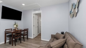 A modern living room with a brown leather sofa, a dining table with chairs, and a flat-screen TV mounted on the wall.