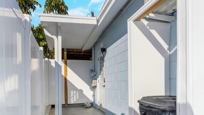A narrow outdoor corridor with a white fence, a black trash bin, and a white wall with a window.