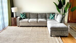A living room with a grey sofa, a small side table with a lamp, a large potted plant, and a view of a door.