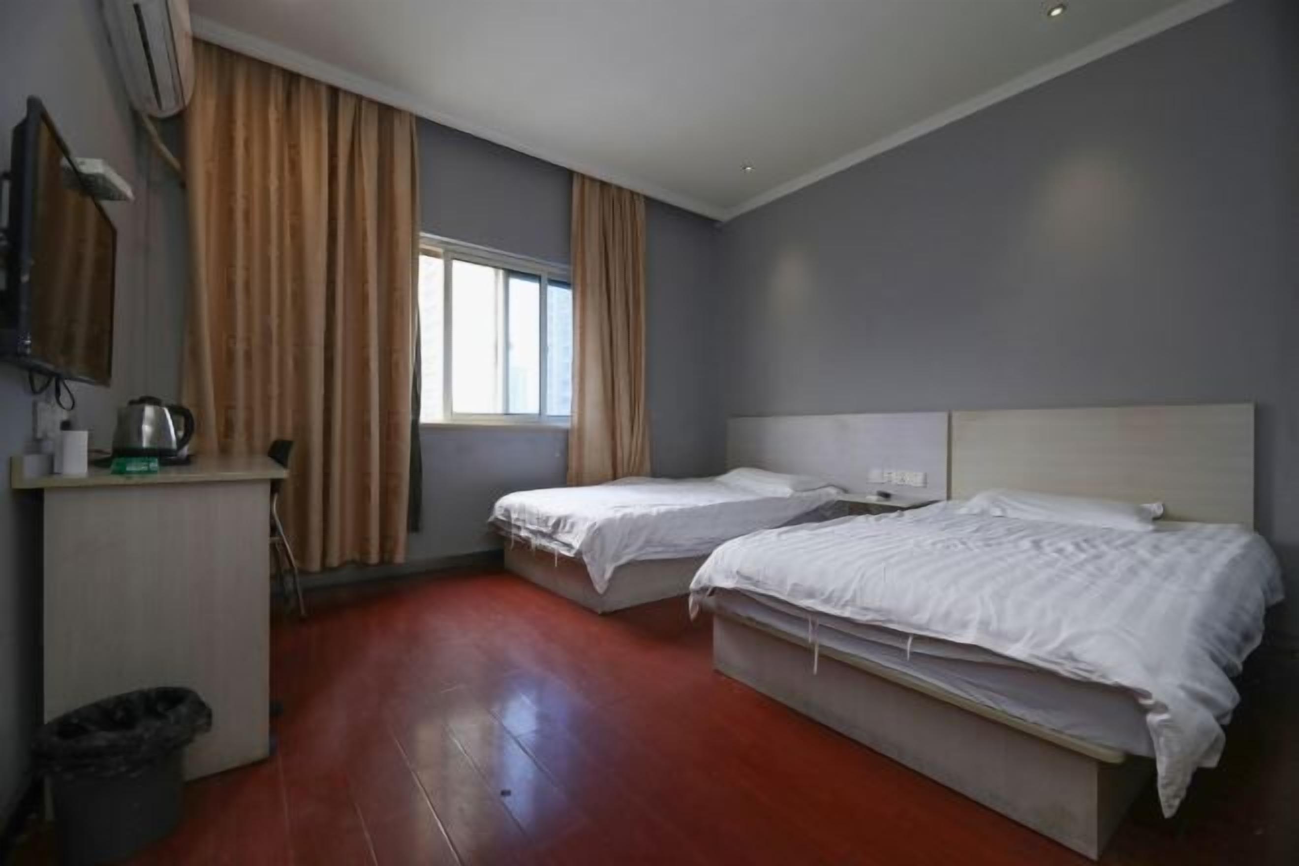 A hotel room with two single beds, a desk with a kettle, a trash bin, and a window with curtains.