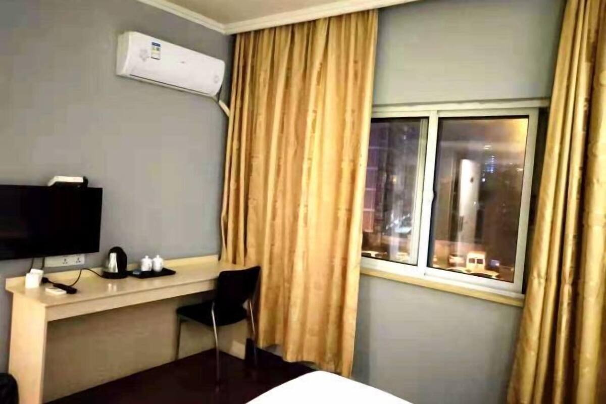 A hotel room with a bed, a desk with a computer, a chair, an air conditioning unit, and a window with curtains.