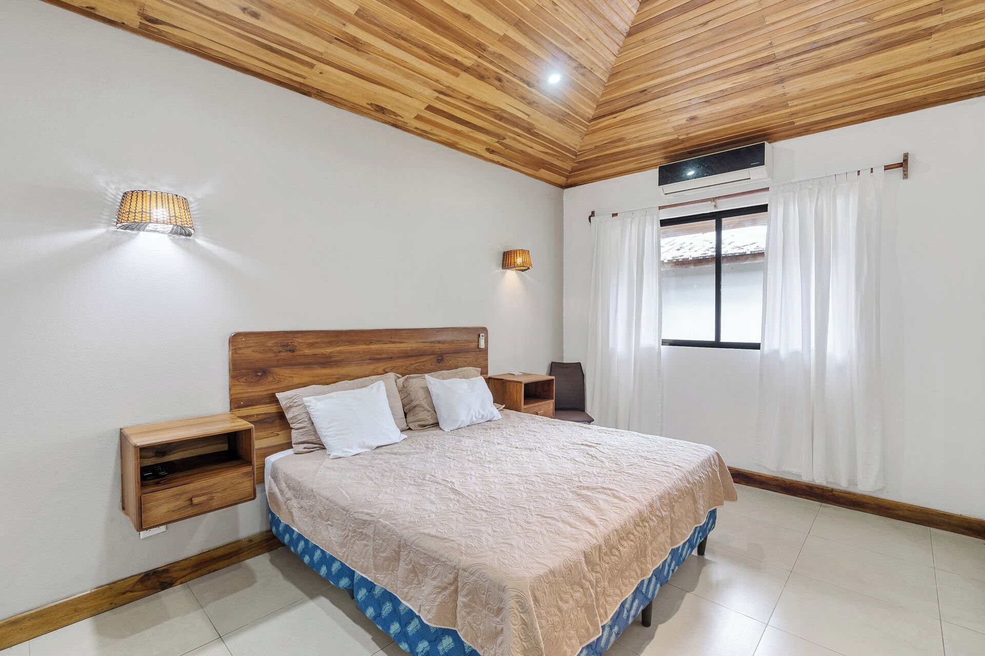 Un dormitorio con techo de madera, cama con colcha estampada, mesitas de noche y una ventana con cortinas.