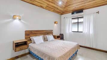 Un dormitorio con techo de madera, cama con colcha estampada, mesitas de noche y una ventana con cortinas.