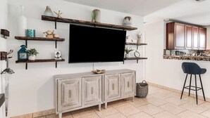 A living room with a TV mounted on the wall, wooden shelves with decorative items, a grey cabinet, and a kitchen area with wooden cabinets and a tiled backsplash. - Relax & Recharge: Saltwater Pool + Lush Backyard Oasis in Palm Beach Gardens (Palm Beach Gardens)
