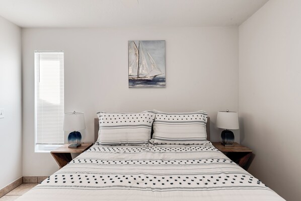 A bedroom with a bed, two bedside tables, a window with blinds, and a painting of a sailboat on the wall.
