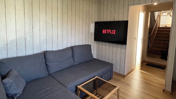 A modern living room with a sectional sofa, a glass coffee table, and a mounted TV displaying 'NETFLIX'. - Cozy 1-room apartment (Hannover)