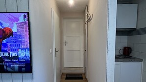 A narrow hallway with a wooden floor leading to a white door. A television mounted on the wall displays a movie. A small kitchen area is visible to the right with a sink and a kettle. - Cozy 1-room apartment (Hannover)