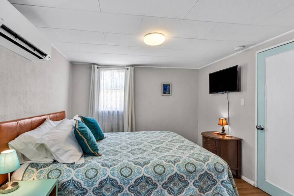 A bedroom with a bed, a bedside table with a lamp, a TV mounted on the wall, and a door.