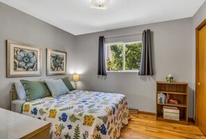 A bedroom with a floral bedspread, a bedside table, a lamp, a window with curtains, and a bookshelf. - Harrison Park Hideaway ~ Dog friendly w. fee (Cañon City)