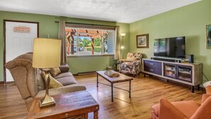 A living room with a sofa, armchair, coffee table, TV stand, and a window with a view of greenery.