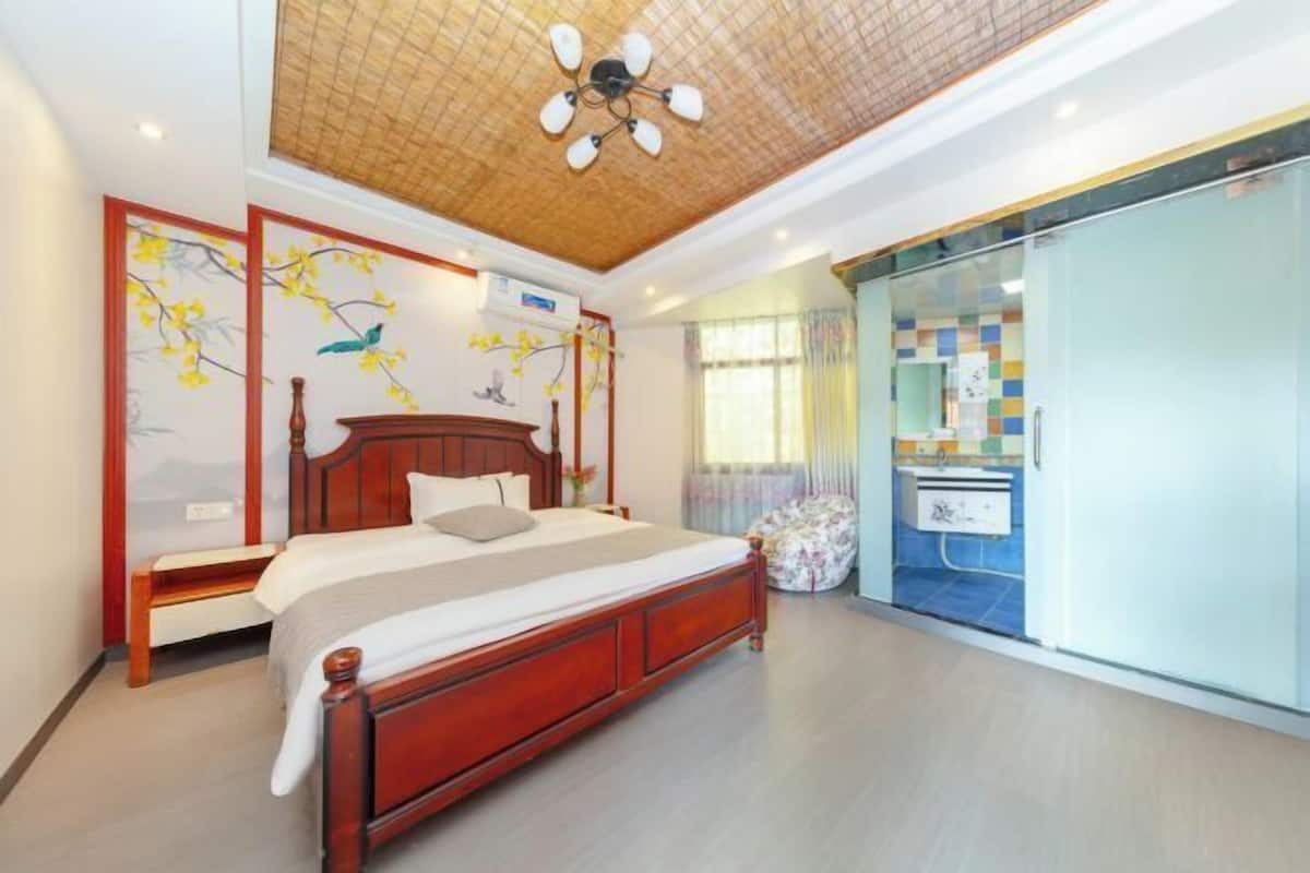 Une chambre avec un lit en bois, un ventilateur de plafond, un climatiseur mural et une porte coulissante en verre donnant sur une salle de bain.