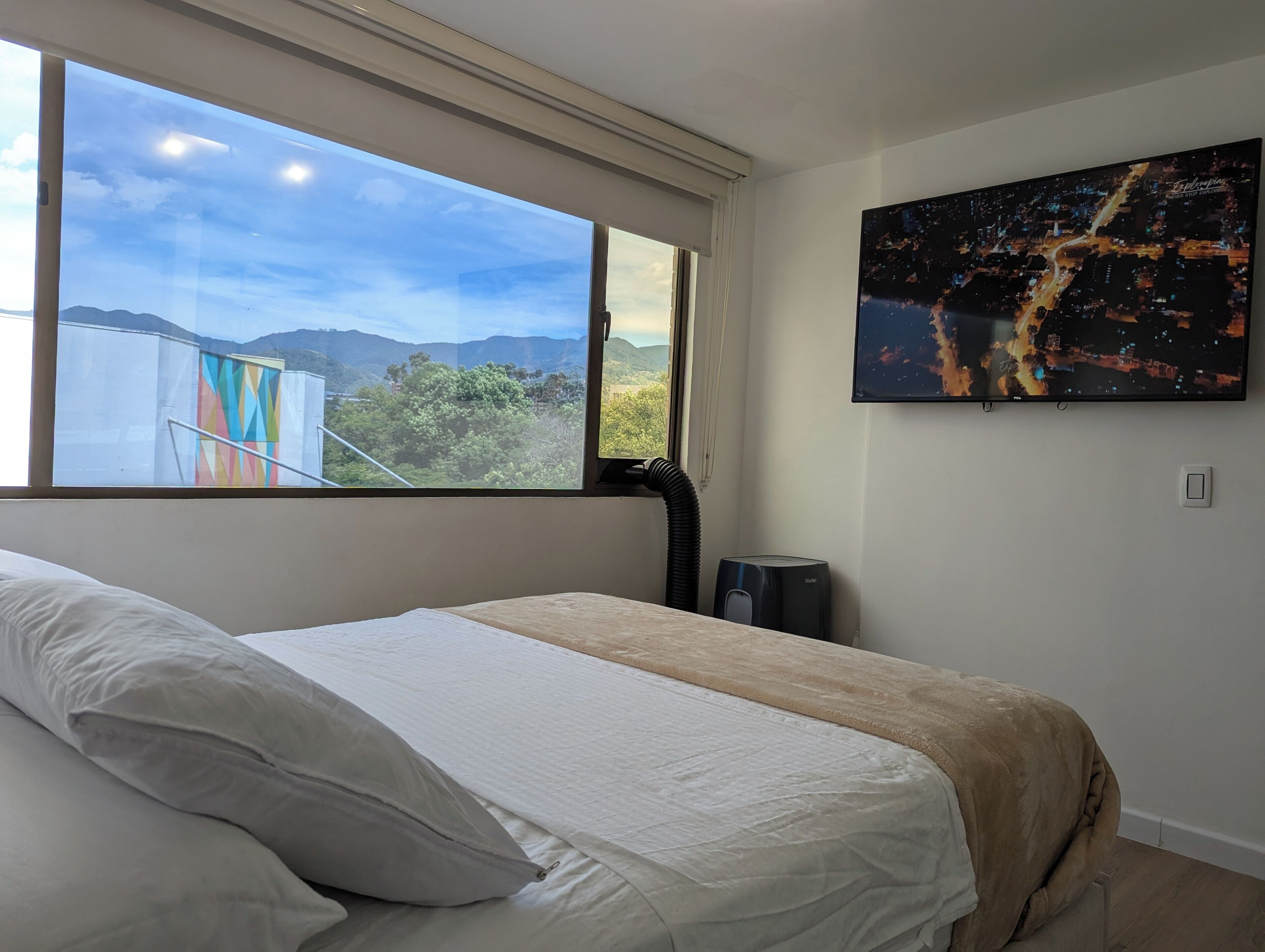 Un dormitorio con un ventanal grande, una cama con ropa de cama blanca, un calefactor y un cuadro de un paisaje urbano en la pared.