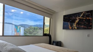 Ein Schlafzimmer mit großem Fenster, einem Bett mit weißer Bettwäsche, einem Heizkörper und einem Stadtbild an der Wand.