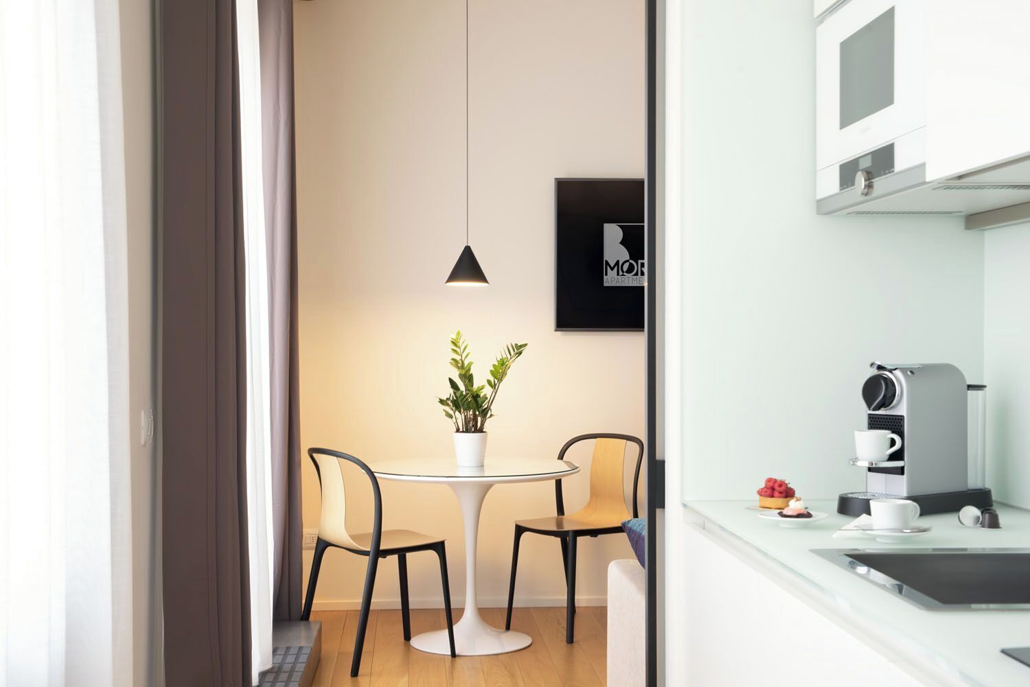 A modern kitchen with a coffee machine, a dining table with chairs, and a potted plant.