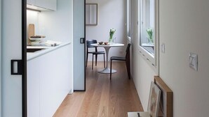 Un pasillo estrecho con una cocina a un lado y un comedor al otro. El suelo es de madera, las paredes son blancas y una ventana proporciona luz natural.