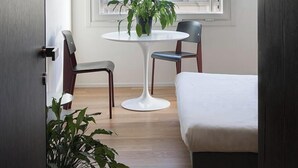 Un dormitorio moderno con una cama, una mesita redonda con una planta, dos sillas y un ventanal que ofrece vistas a edificios.