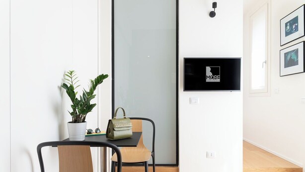 A modern room with a wooden floor, a black-framed desk with a potted plant, a green handbag, and a wall-mounted TV displaying "MORE APARTMENTS". - Penthouse Apartment 5.4 (Milano)