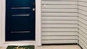 Une porte bleu foncé avec un cadre blanc, un petit luminaire au-dessus, et un paillasson vert sur lequel est écrit le mot « Joie ».