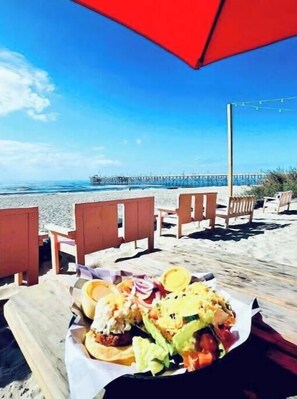 Escena a la vora de la platja amb menjar, cadires de fusta i un parasol vermell.