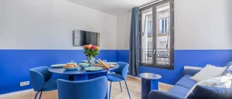 A modern living room with a blue and white color scheme, a dining table with chairs, a sofa, and a TV mounted on the wall.
