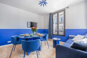A modern living room with a blue and white color scheme, a dining table with chairs, a sofa, and a TV mounted on the wall. - 10min from Paris | Close to Shops/ 8min from Metro (Boulogne-Billancourt)