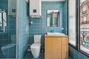 Une salle de bain avec des carreaux bleus, des toilettes blanches, un meuble en bois et une fenêtre donnant sur des immeubles.