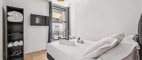 A bedroom with a bed, a shelf with towels, a TV, and a window with a balcony.