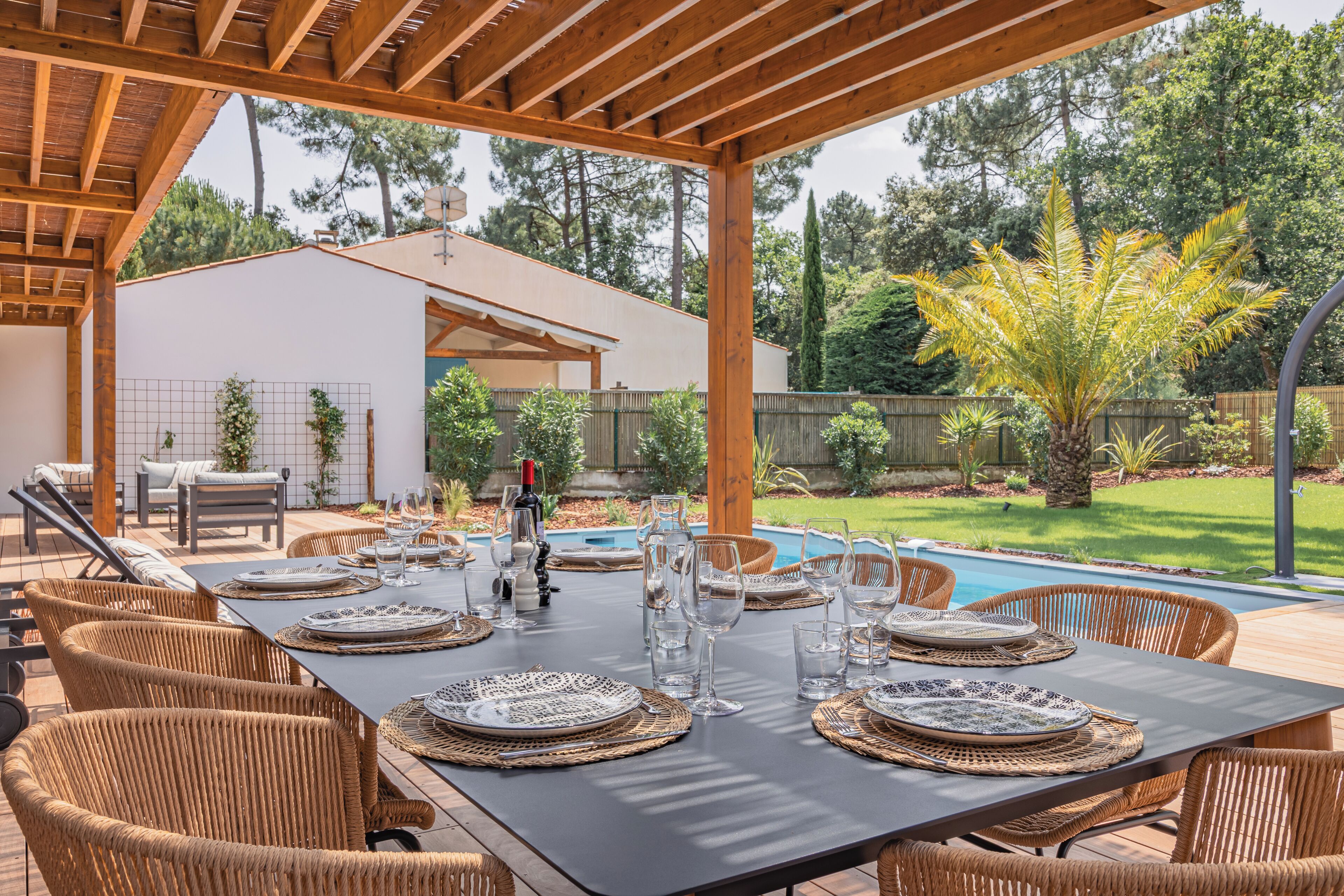 Uma área de refeições ao ar livre com uma mesa posta para seis pessoas, rodeada por cadeiras de vime, sob uma pérgola de madeira. Uma piscina é visível ao fundo.