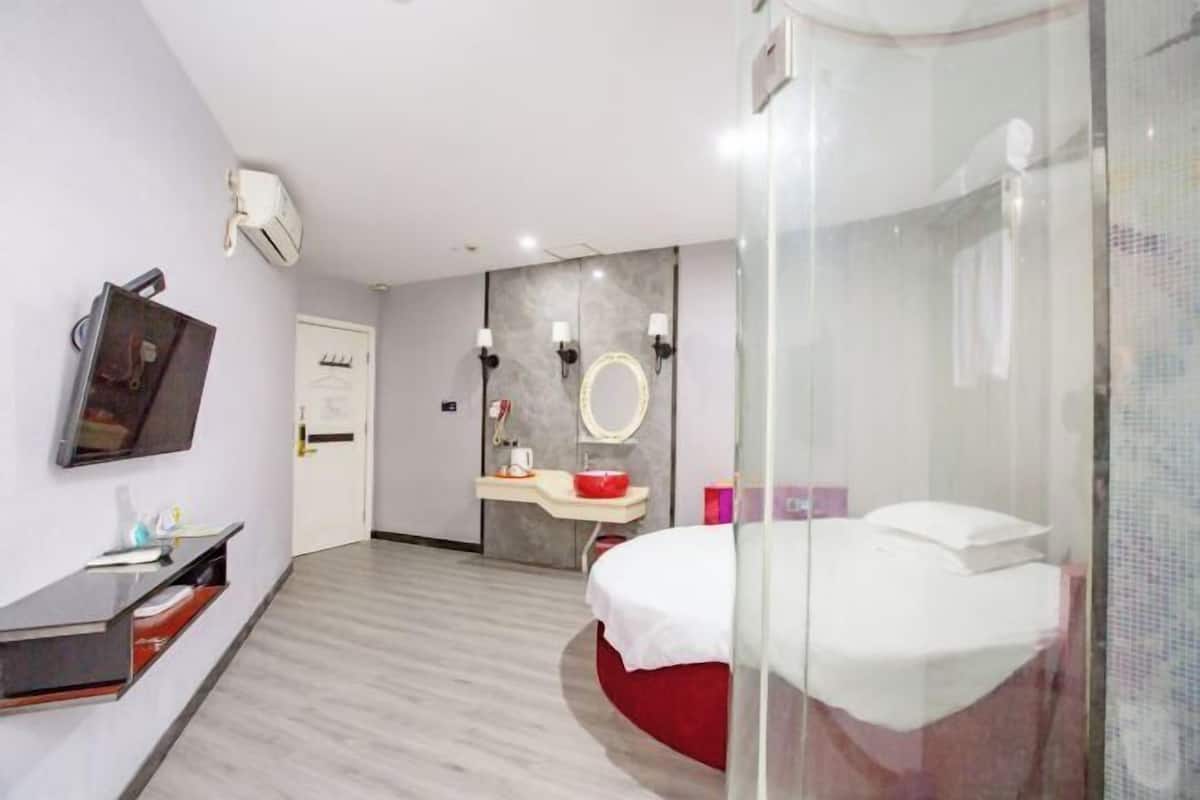 A modern hotel room with a bed, a desk with a flat-screen TV, a vanity with a mirror, and a shower area.