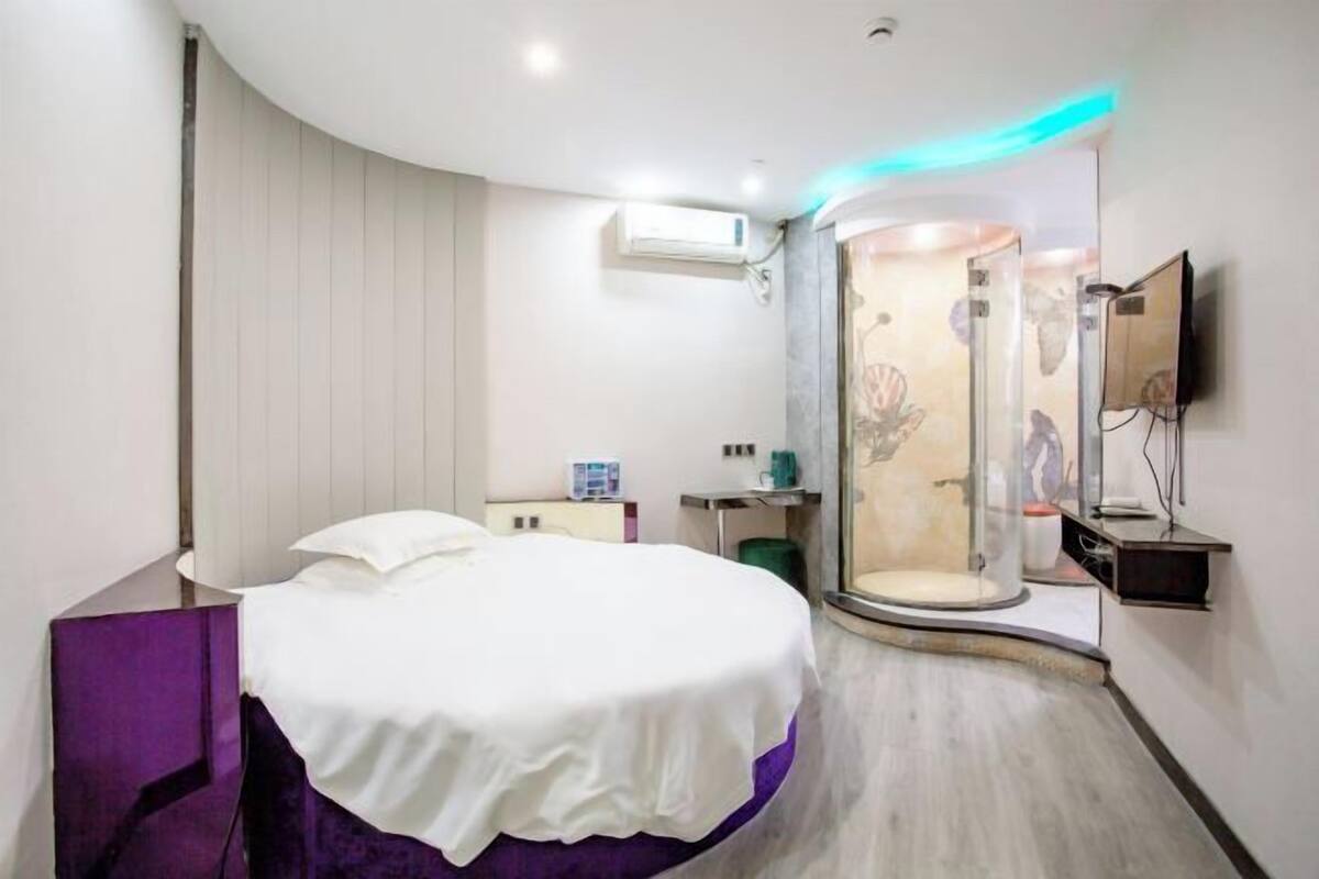A modern hotel room with a curved bed, a shower enclosure, a mounted TV, and a small desk with a chair.