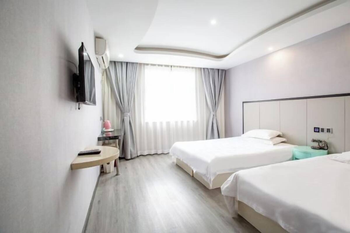 A hotel room with two beds, a TV mounted on the wall, a bedside table, a window with curtains, and a modern ceiling design.