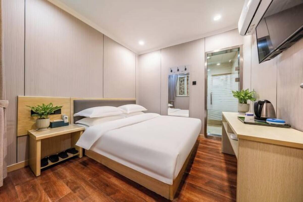Une chambre d’hôtel moderne avec un plancher en bois, un lit avec une literie blanche, une table de chevet agrémentée d’une plante, un bureau avec une bouilloire et un téléviseur fixé au mur.