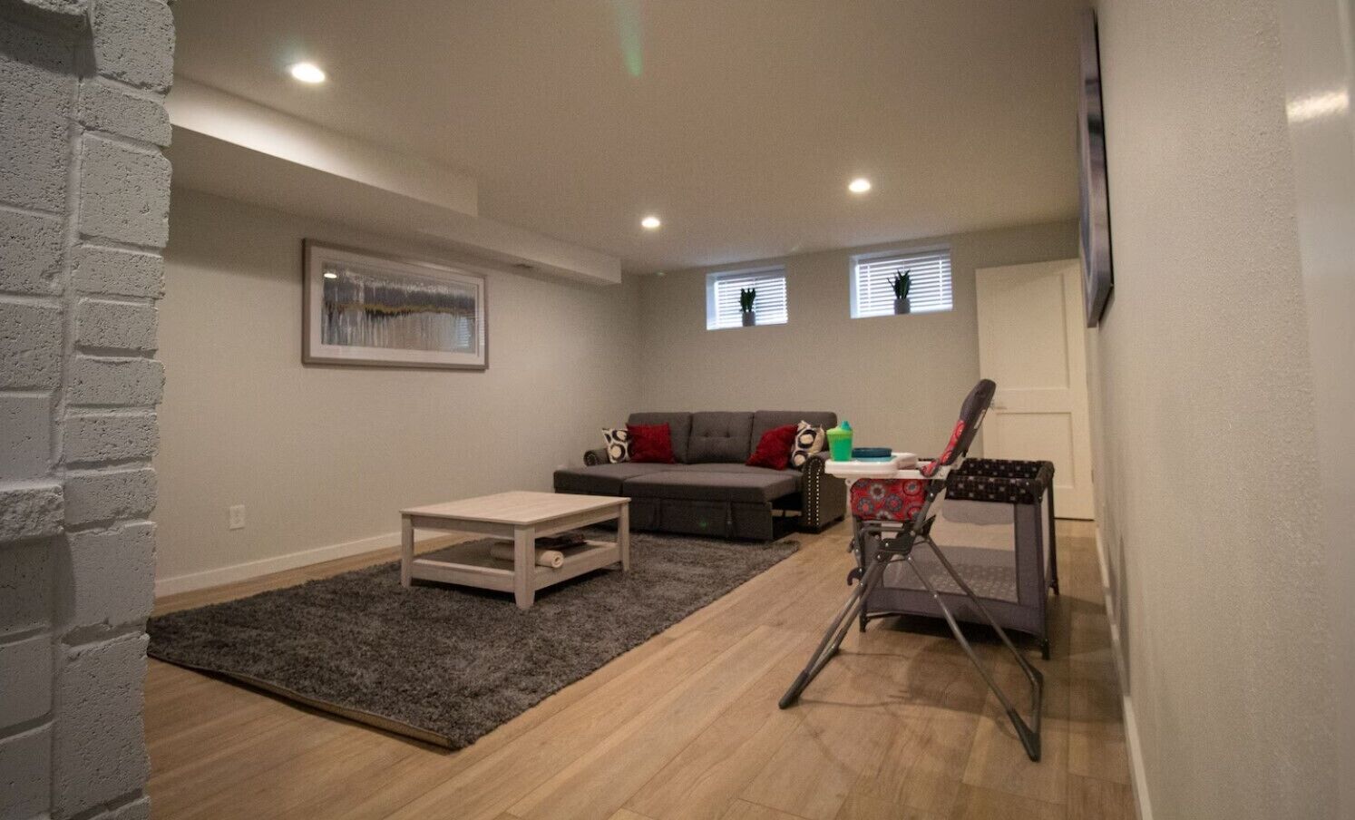Un salon avec un canapé gris, une petite table et un transat pour bébé. Il y a deux fenêtres avec des stores, un tableau encadré au mur et un tapis gris.